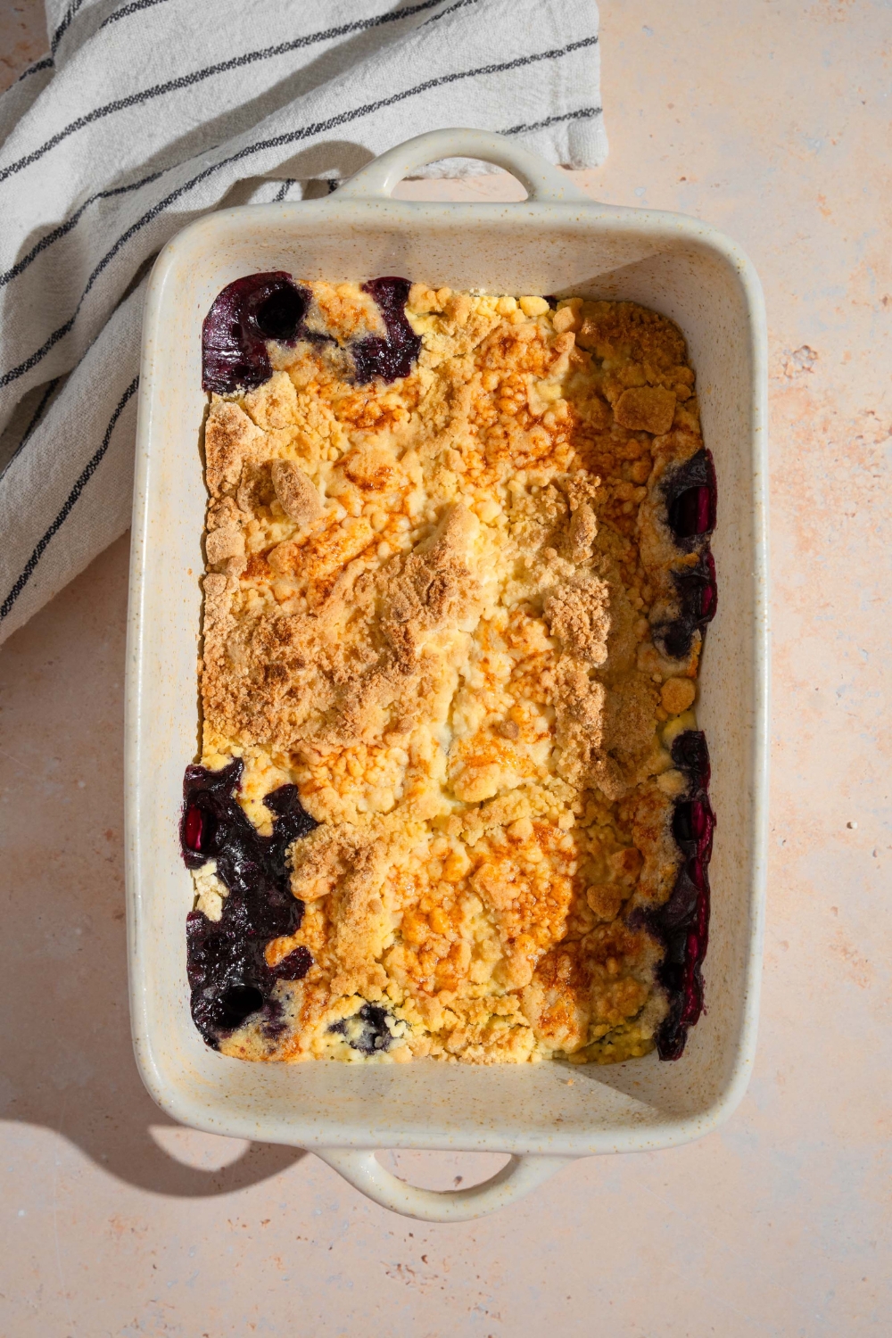 A baking dish with baked blueberry dump cake. The baking dish is on a tan counter with a white striped napkin.