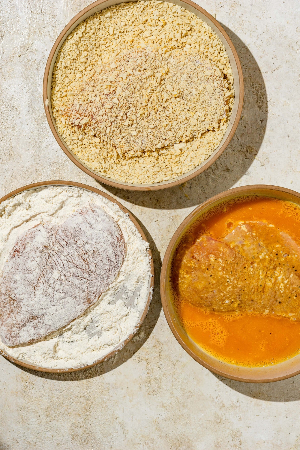 A plate of flour mixture, a plate of panko breadcrumbs, and a bowl of beaten eggs on a tan counter. There is an uncooked chicken breast in each plate.