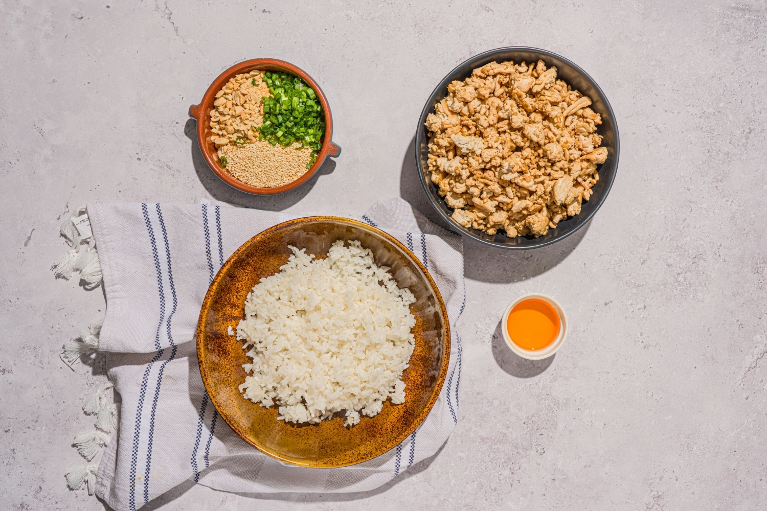 A white counter with three bowls containing ingredients to make ground turkey bowls. One bowl has cooked rice. A second bowl has cooked seasoned ground turkey. The third bowl has toppings including sesame seeds, peanuts, and sliced green onions.