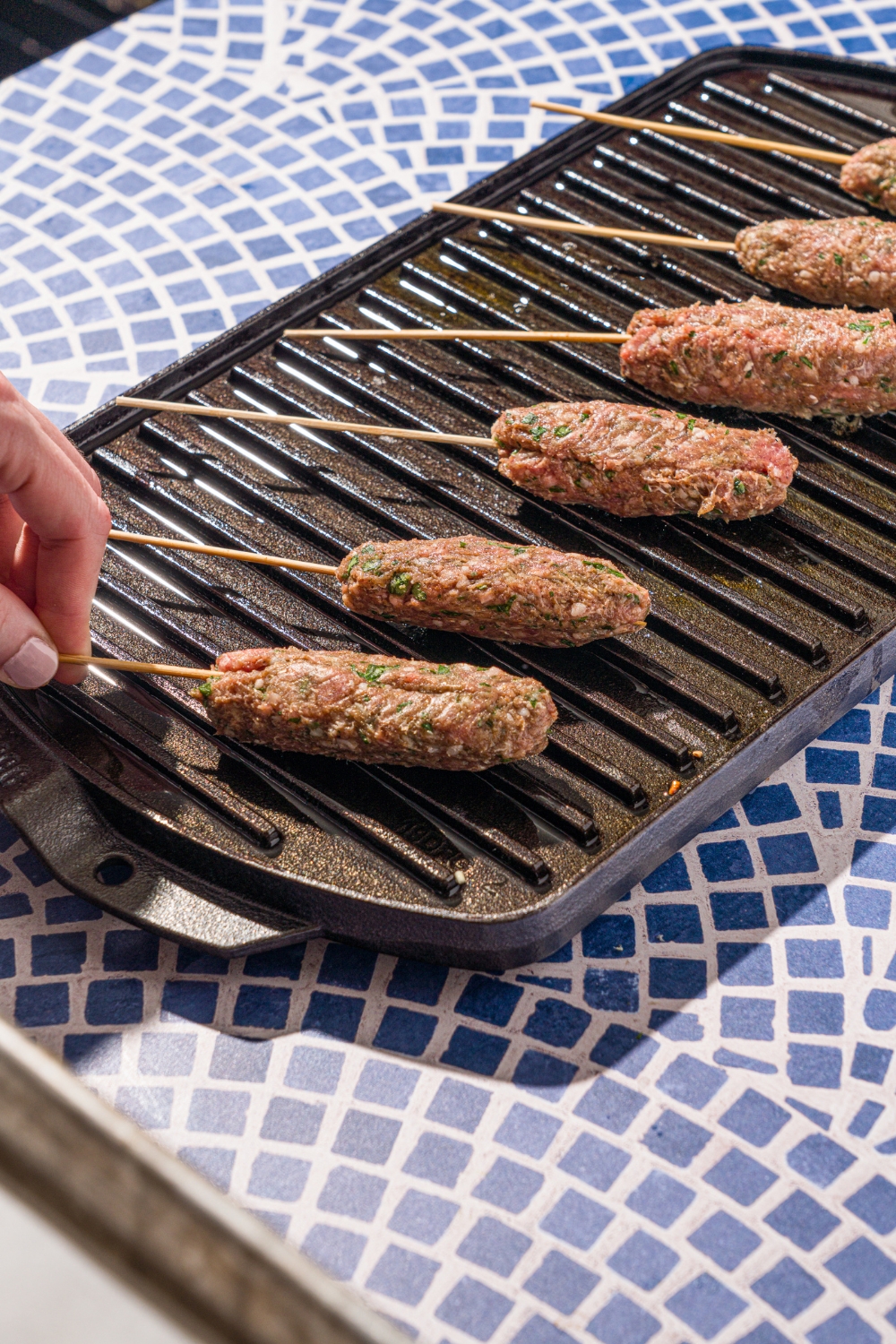 An indoor grill with several Kafta kabobs grilling. A hand is placing a kabob on the grill. The grill is on a tiled counter.