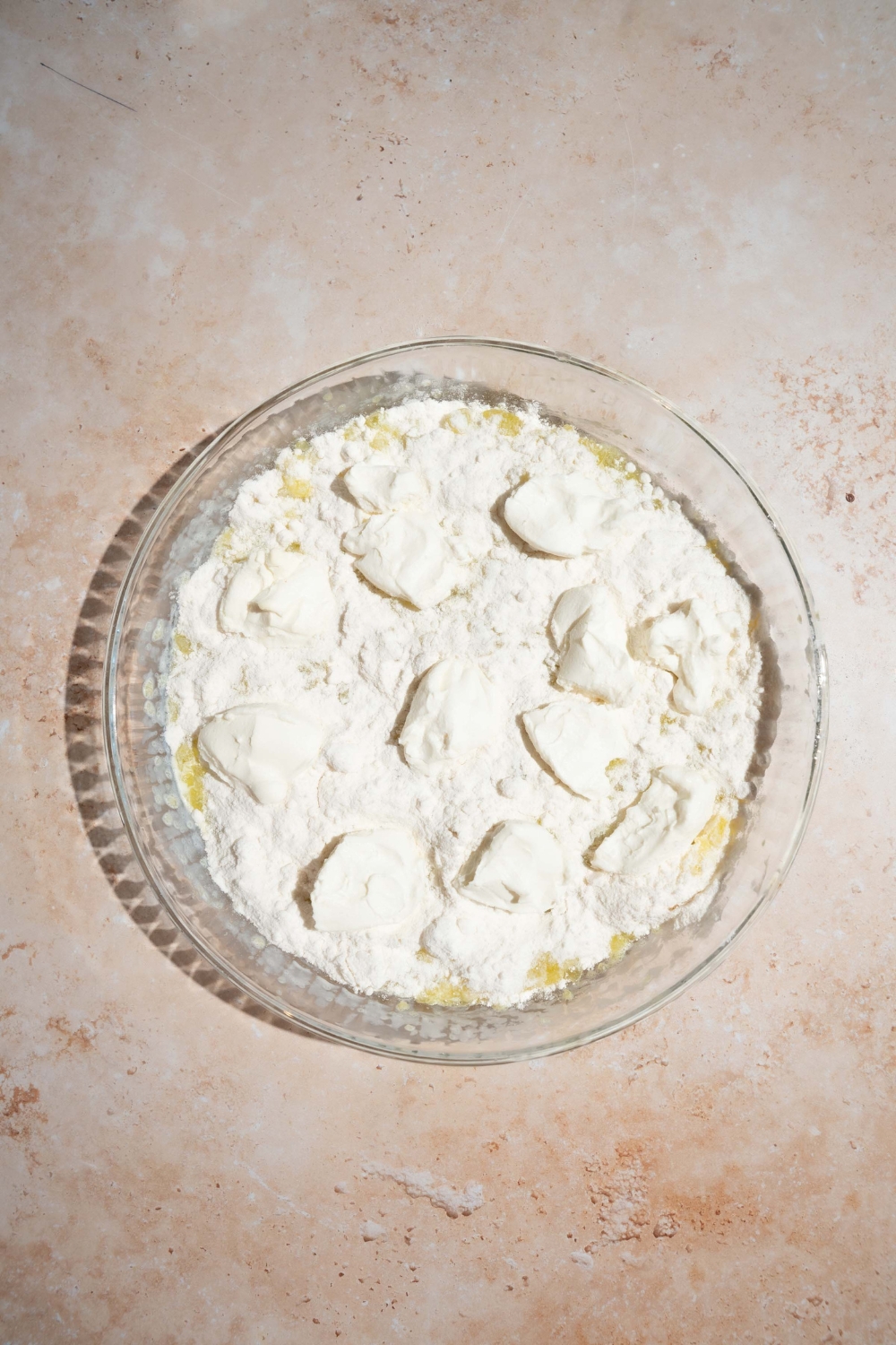 A glass baking dish with layered ingredients to make lemon cream cheese dump cake including lemon filling, cake mix, and cream cheese. The dish is on a tan counter.