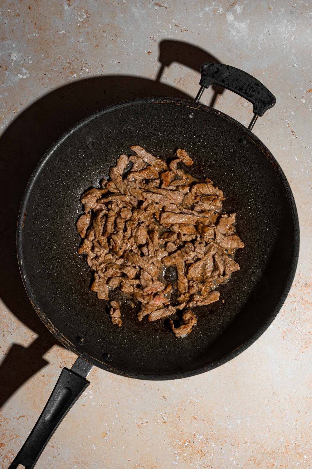 A wok with sliced steak cooking in oil. The wok is on a tan counter.