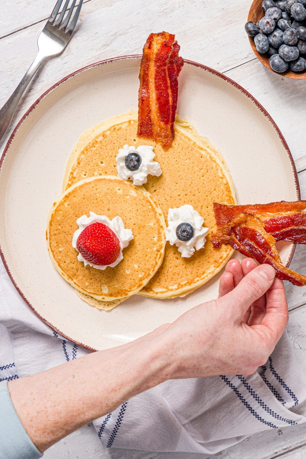 A white plate with reindeer pancakes topped with whipped cream, blueberries, and a strawberry. A hand is adding bacon strips to the pancakes. The plate is on a wood counter with a bowl of blueberries.
