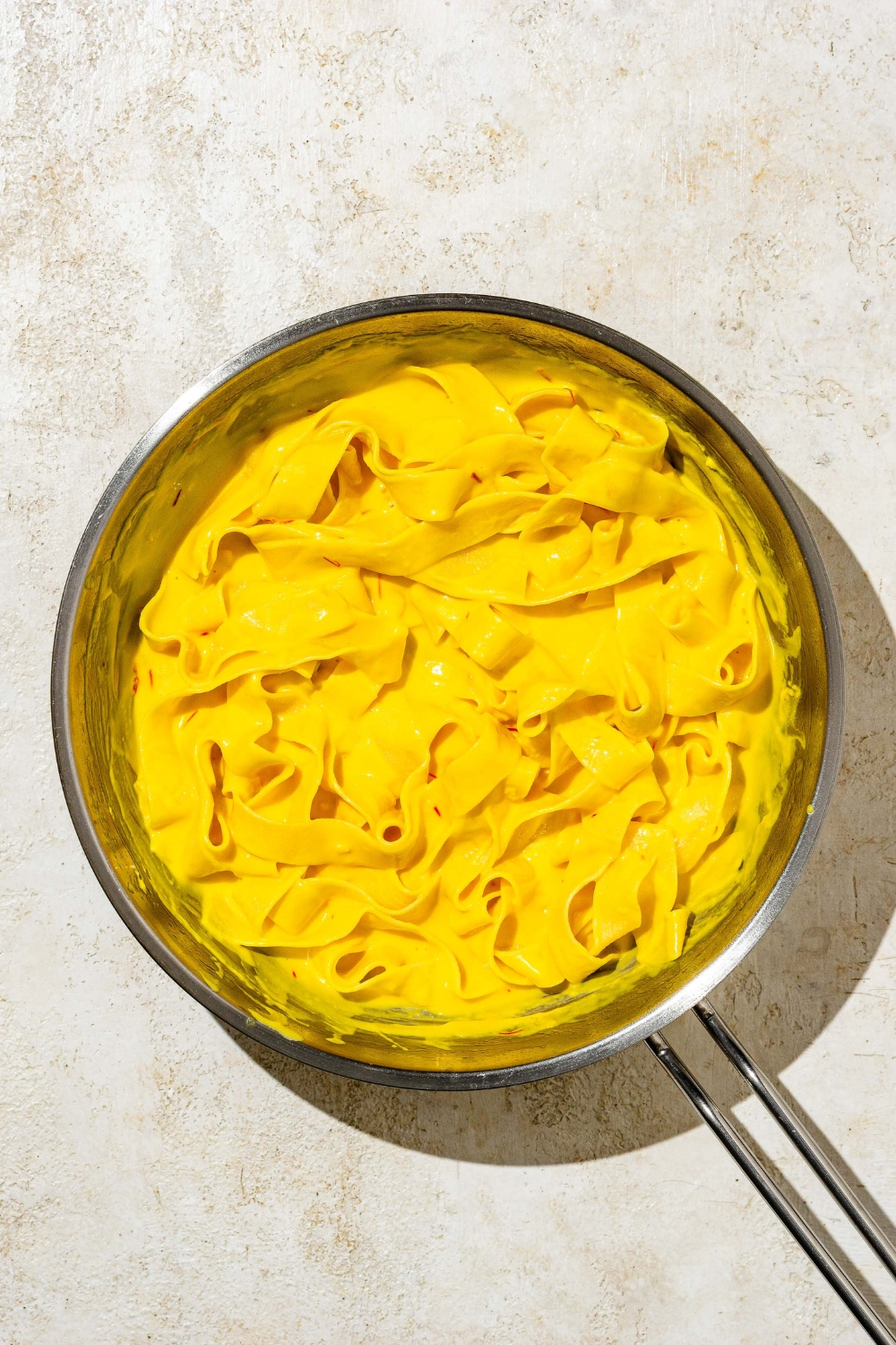 A skillet with fettuccine pasta tossed in saffron cream sauce. The skillet is on a tan counter.