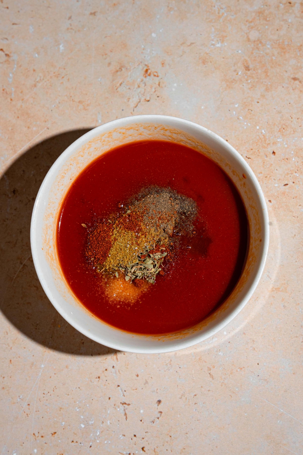 A glass bowl with ingredients to make taco sauce such as tomato sauce and spices. The bowl is on a tan counter.