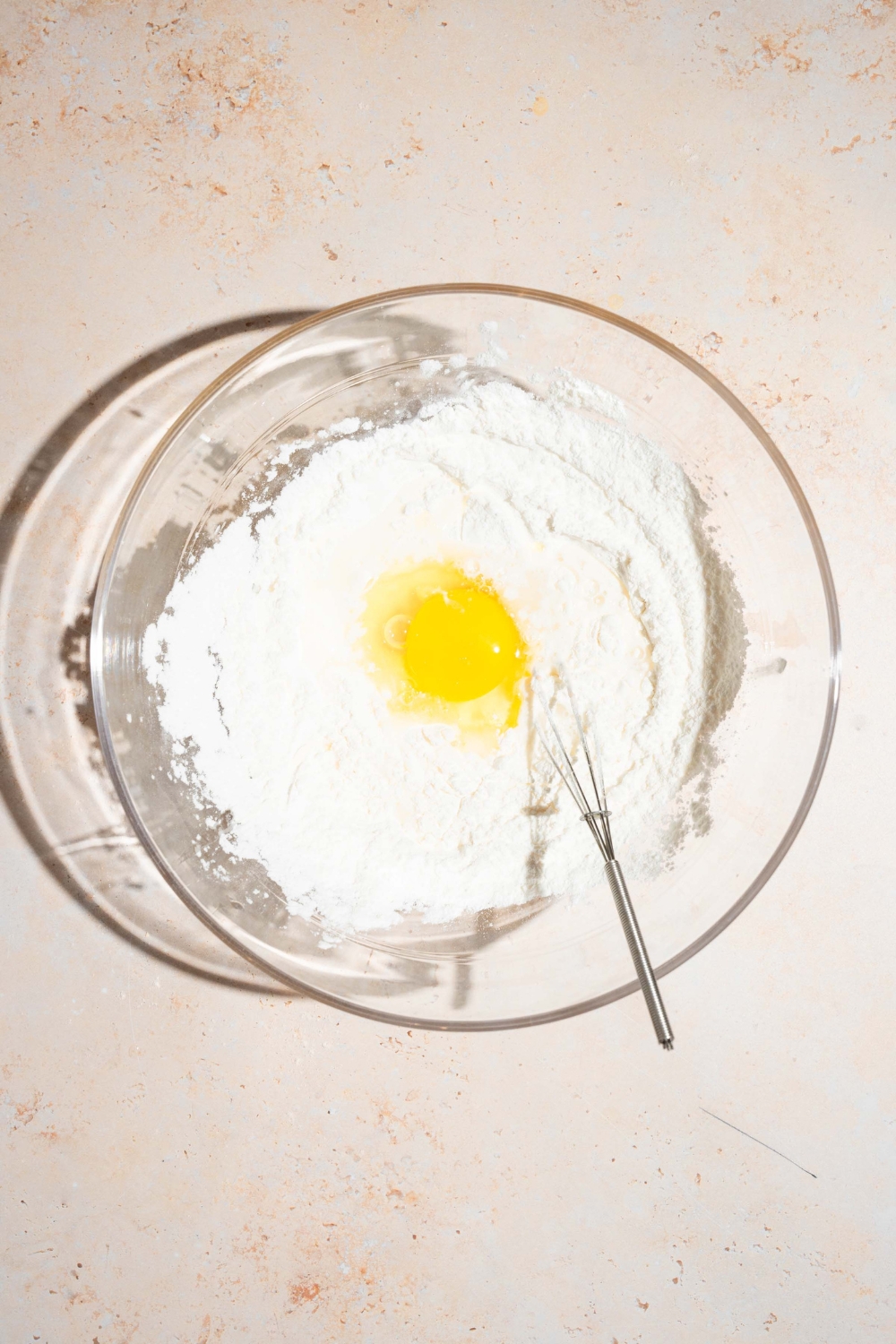 A glass bowl with a whisk mixing dry ingredients with an egg. The bowl is on a tan counter.