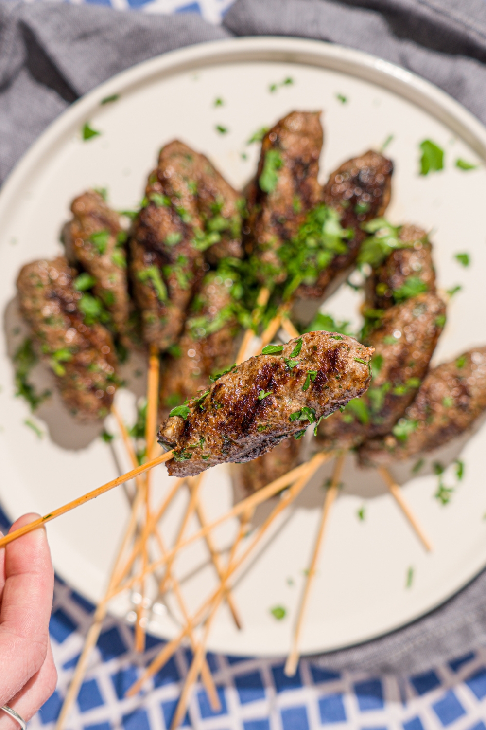 A close up of a hand holding a Kafta kabob garnished with fresh parsley. There is a white plate with additional kabobs blurred in the background.