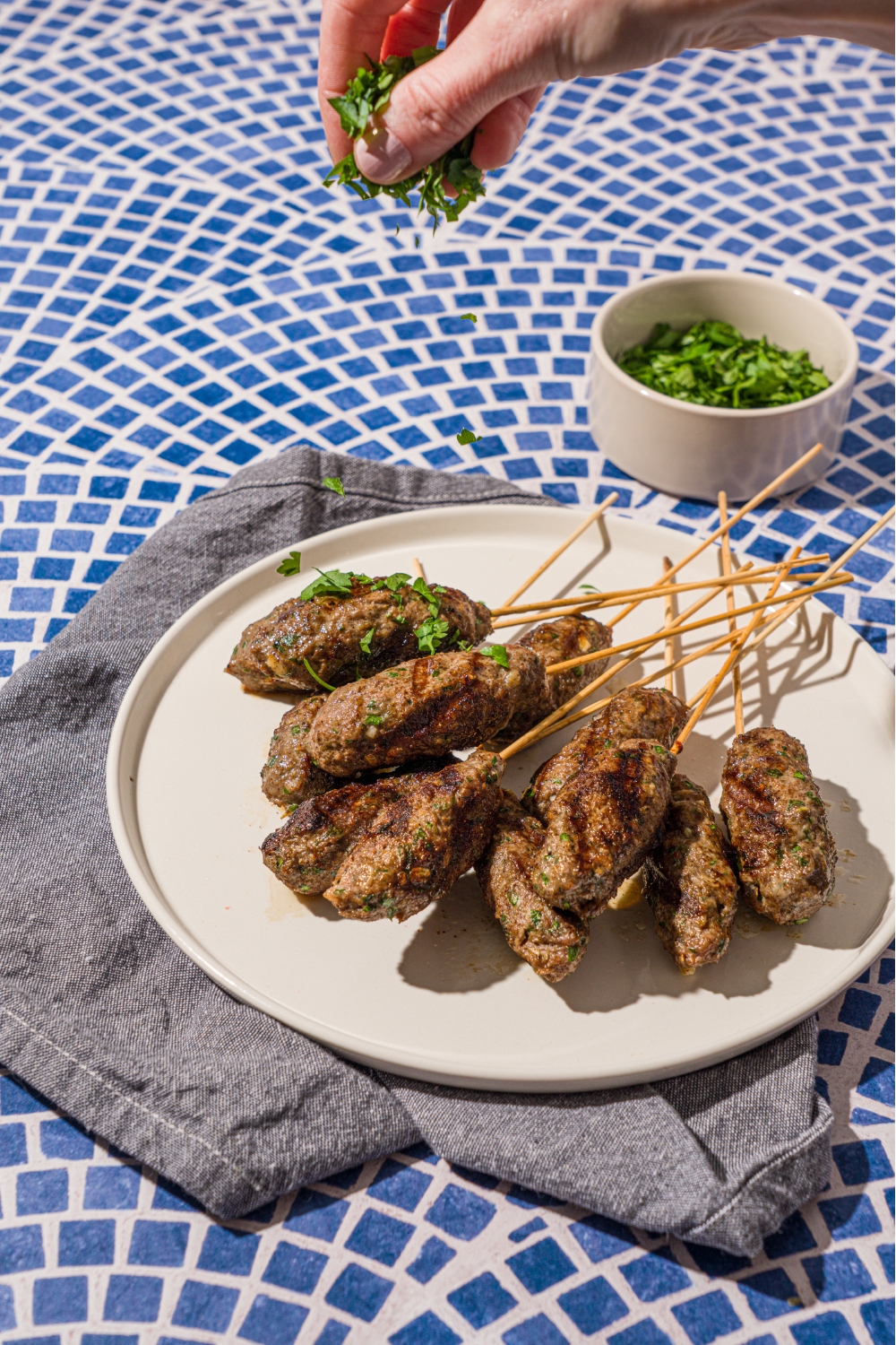 A white plate with a pile of Kafta kabobs. A hand is sprinkling fresh parsley over the kabobs. The plate is on a tiled counter with a gray cloth napkin.