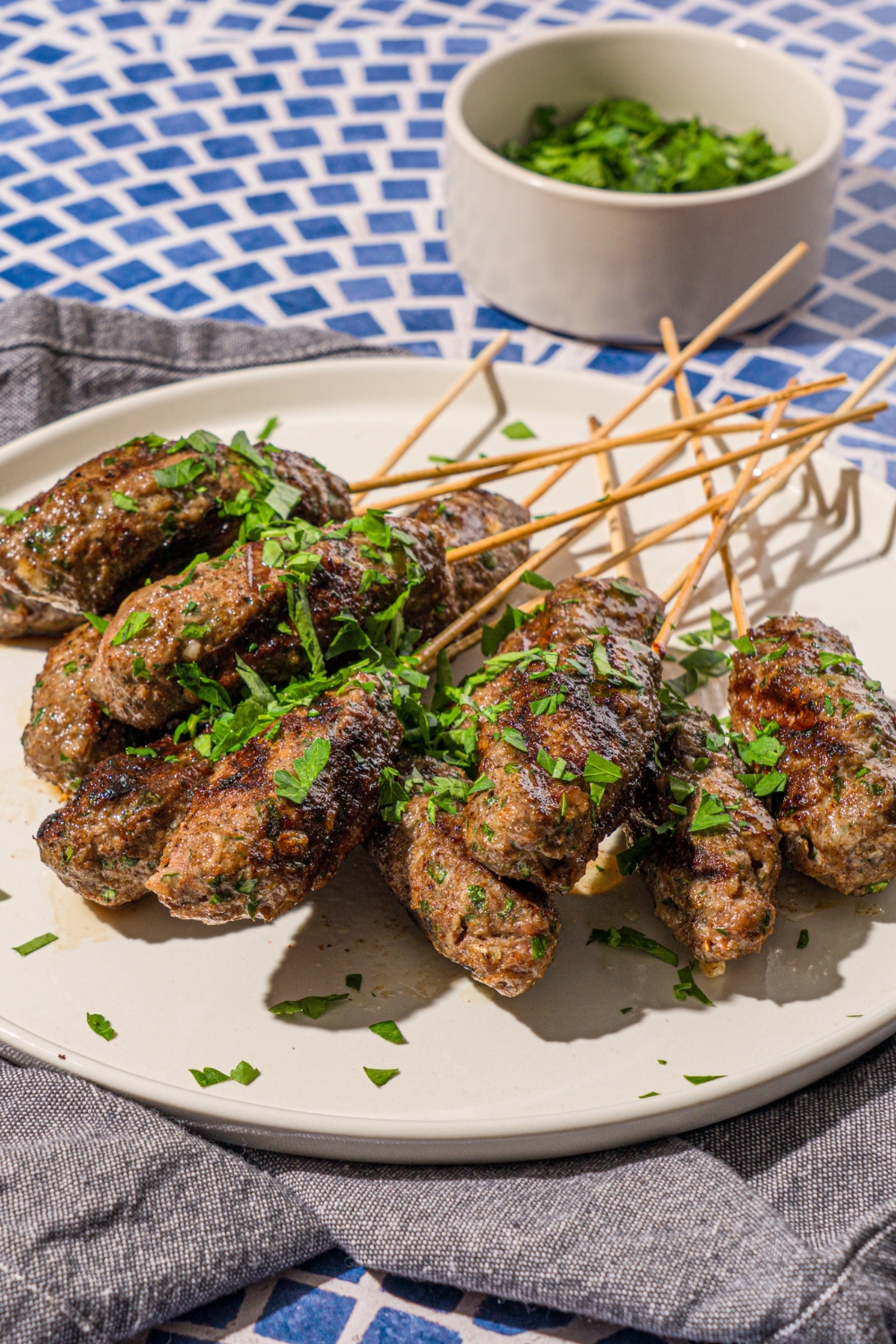 A white plate with a pile of Kafta kabobs garnished with fresh parsley. The plate is on a tiled counter with a gray cloth napkin.