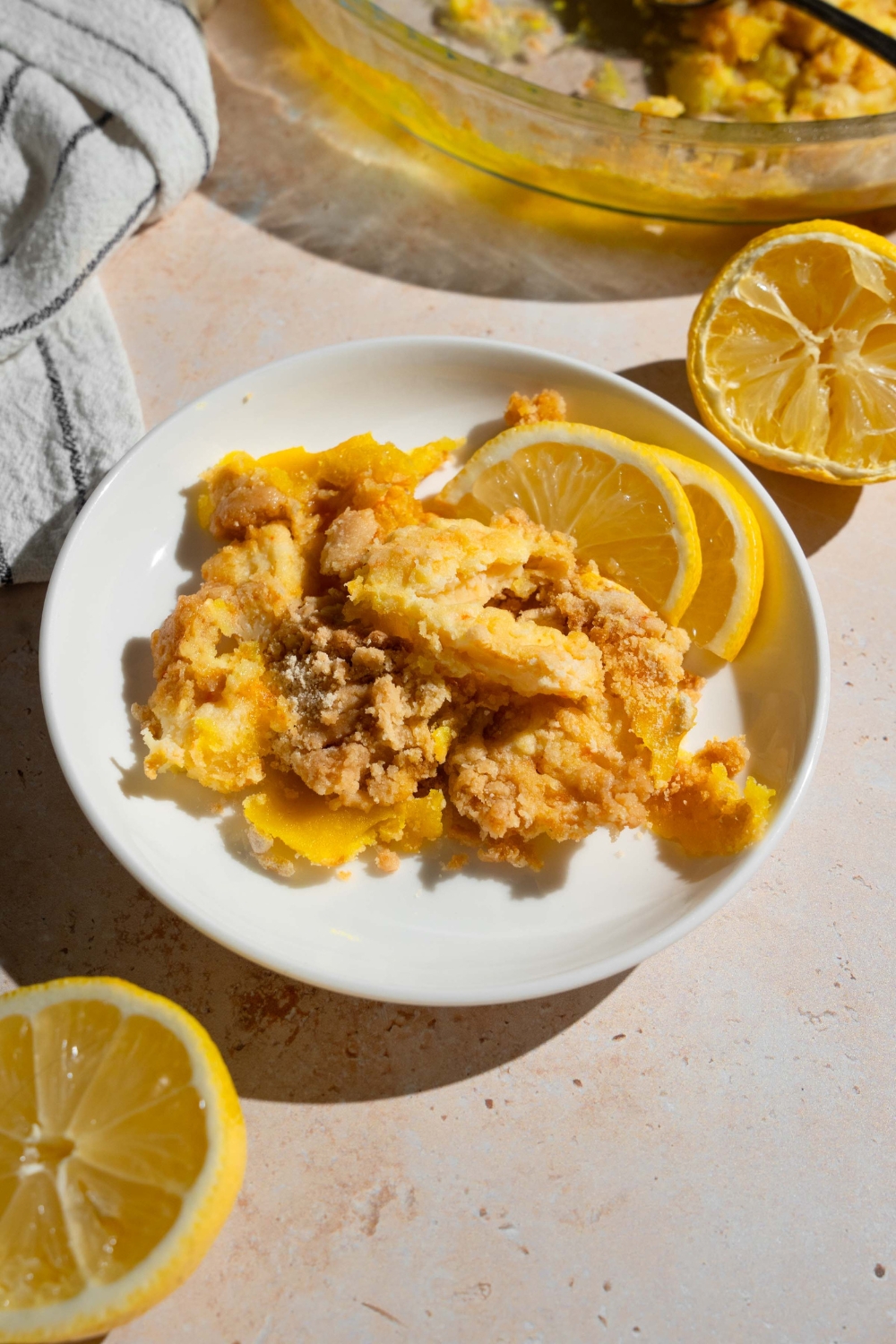 A white plate with a slice of lemon cream cheese dump cake served with sliced lemon. The plate is on a tan counter with a baking dish with cake, lemons, and a white striped napkin.