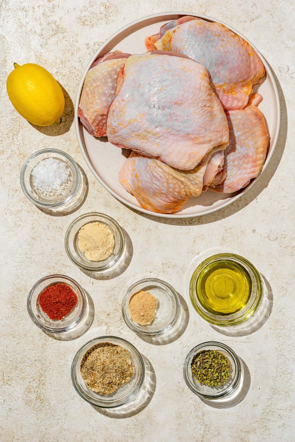 An overhead shot of several bowls in various sizes containing ingredients for lemon pepper chicken thighs including chicken thighs, lemon, oil, lemon pepper seasoning, garlic powder, onion powder, and paprika.