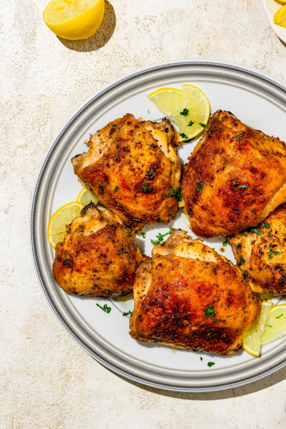 A plate of lemon pepper chicken thighs garnished with fresh parsley and lemon. The plate is on a tan counter with lemon.