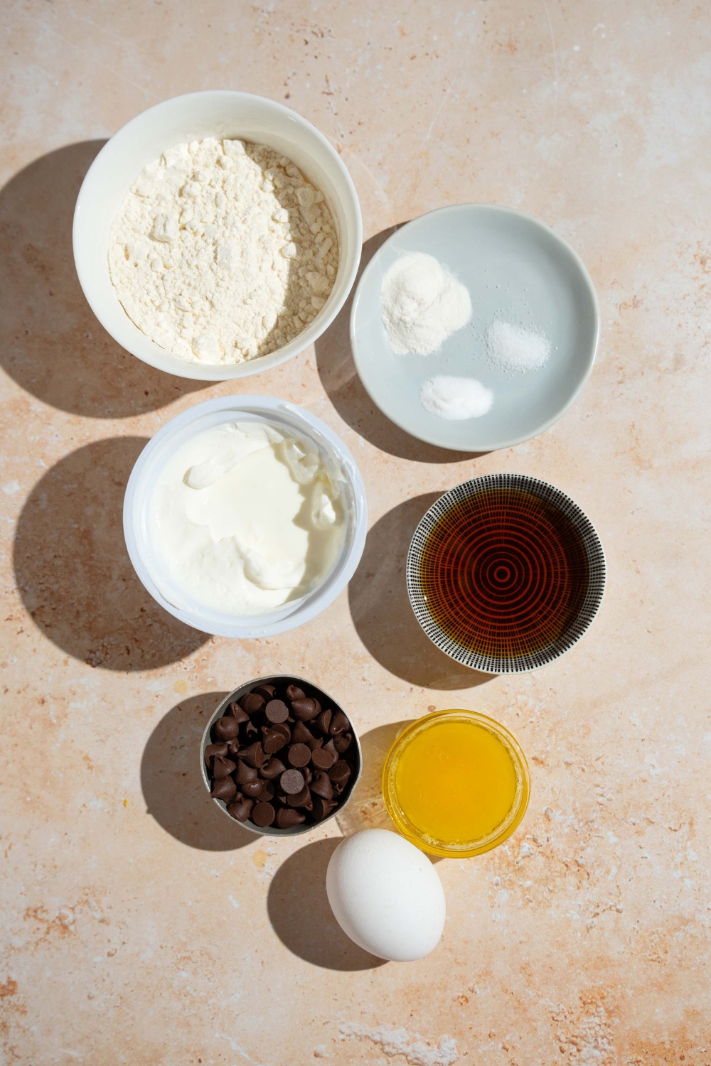 An overhead shot of several bowls in various sizes containing ingredients to make copycat Little Bites muffins including flour, yogurt, eggs, butter, vanilla, baking soda, baking powder, and chocolate chips.