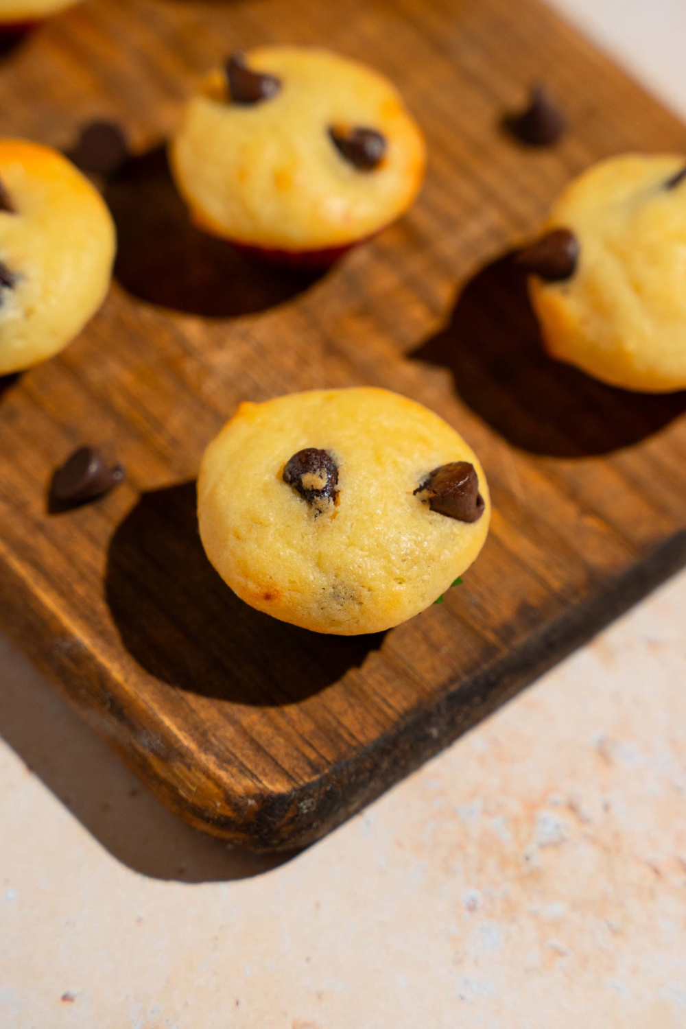 A wooden board with copycat Little Bites chocolate chip muffins. The board is garnished with chocolate chips. The board is on a tan counter.