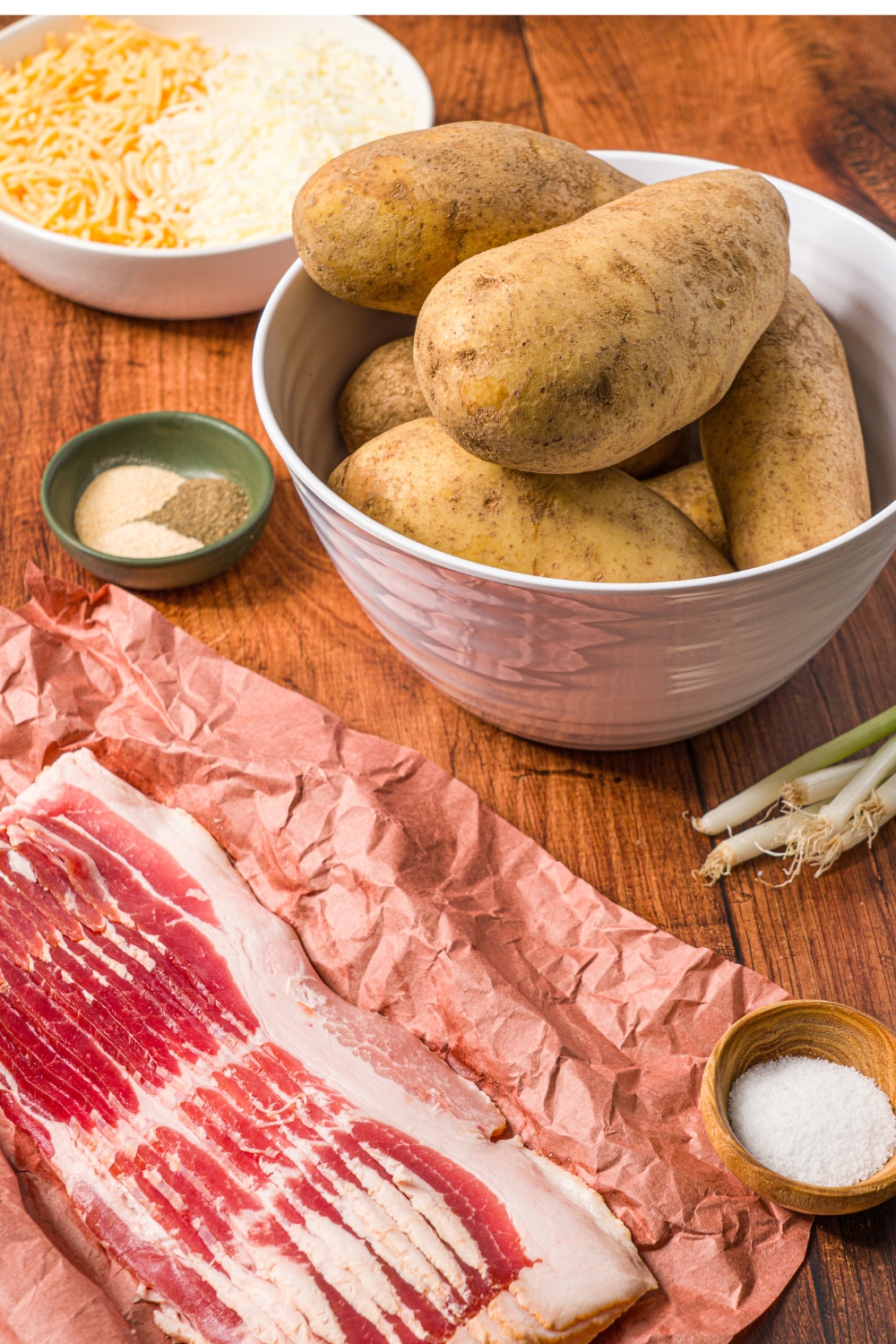 A wooden counter with ingredients to make loaded potato skins including a bowl of potatoes, a slab of uncooked bacon, a bowl of shredded cheeses, and small bowls of seasonings.