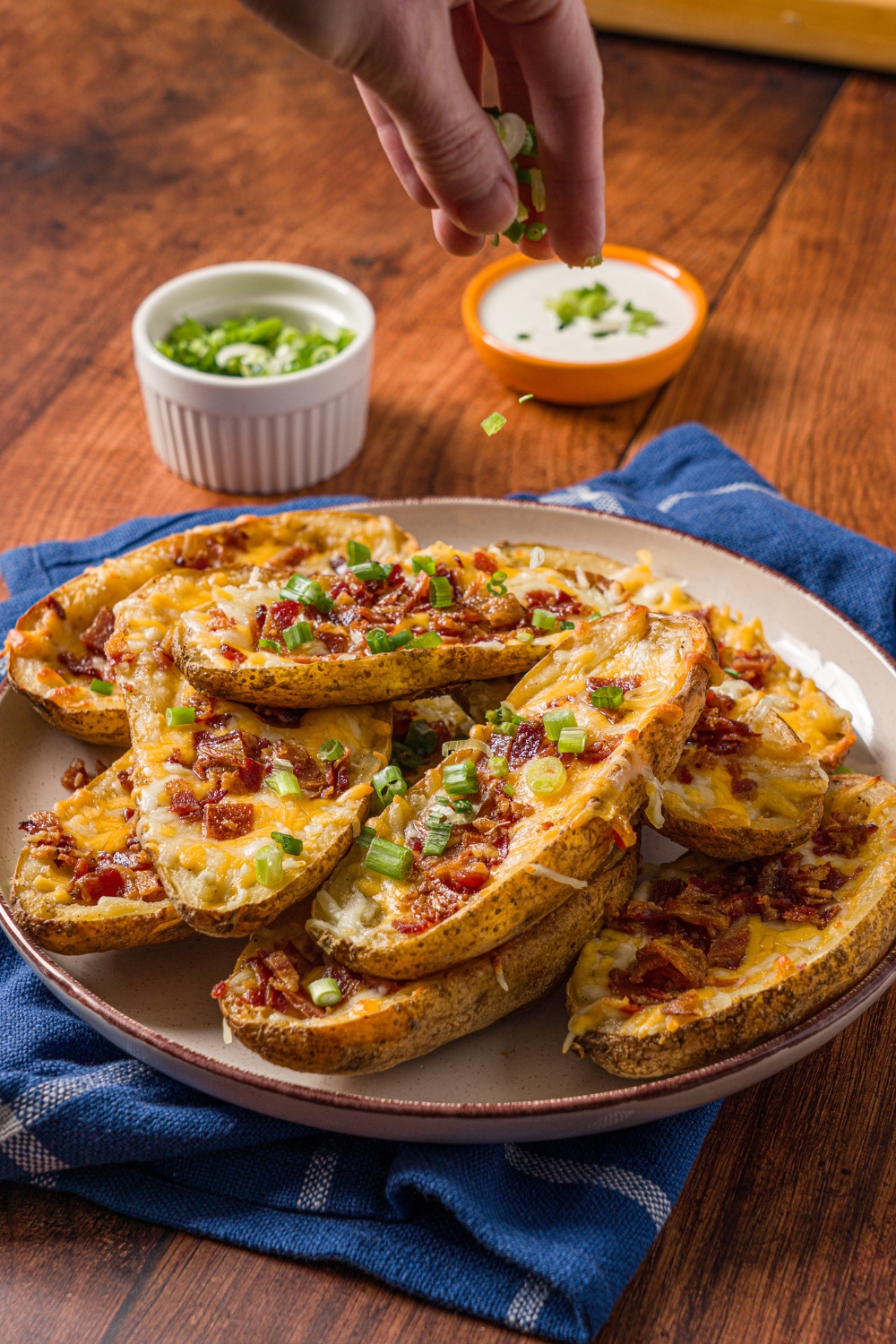 A white plate with a pile of loaded potato skins with a hand sprinkling sliced green onions. The plate is on a wood counter with a blue striped napkin, small bowl of garnishes, and small bowl of dip.