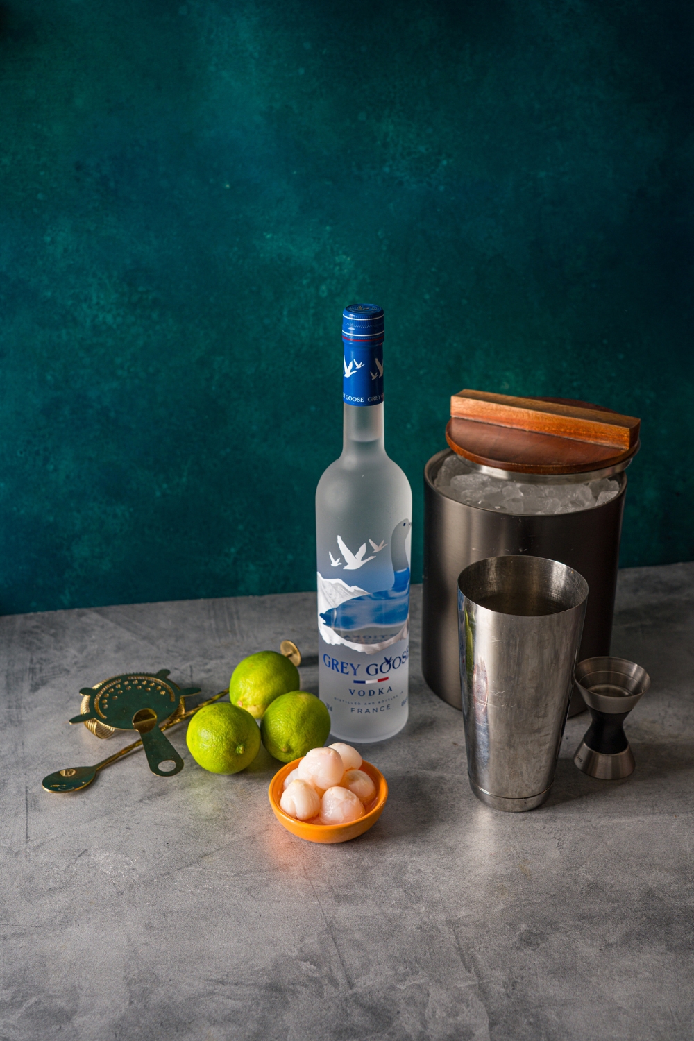 Ingredients to make lychee martini including a bottle of Grey Goose vodka, cocktail shaker, jigger, small bowl of lychees, limes, and an ice bucket.