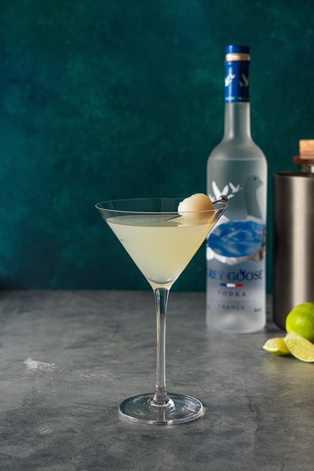 A lychee martini garnished with a lychee in a toothpick. The martini is on a stone counter with a bottle of Grey Goose.