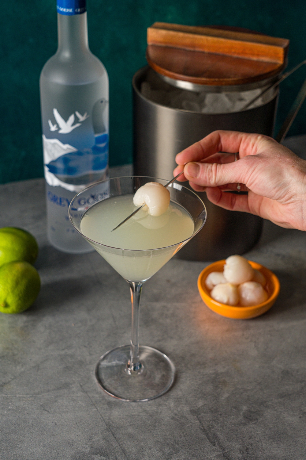 A hand placing a lychee stabbed in a toothpick into a lychee martini. The martini is on a stone counter with a small bowl of lychees, bottle of Grey Goose, and an ice bucket.