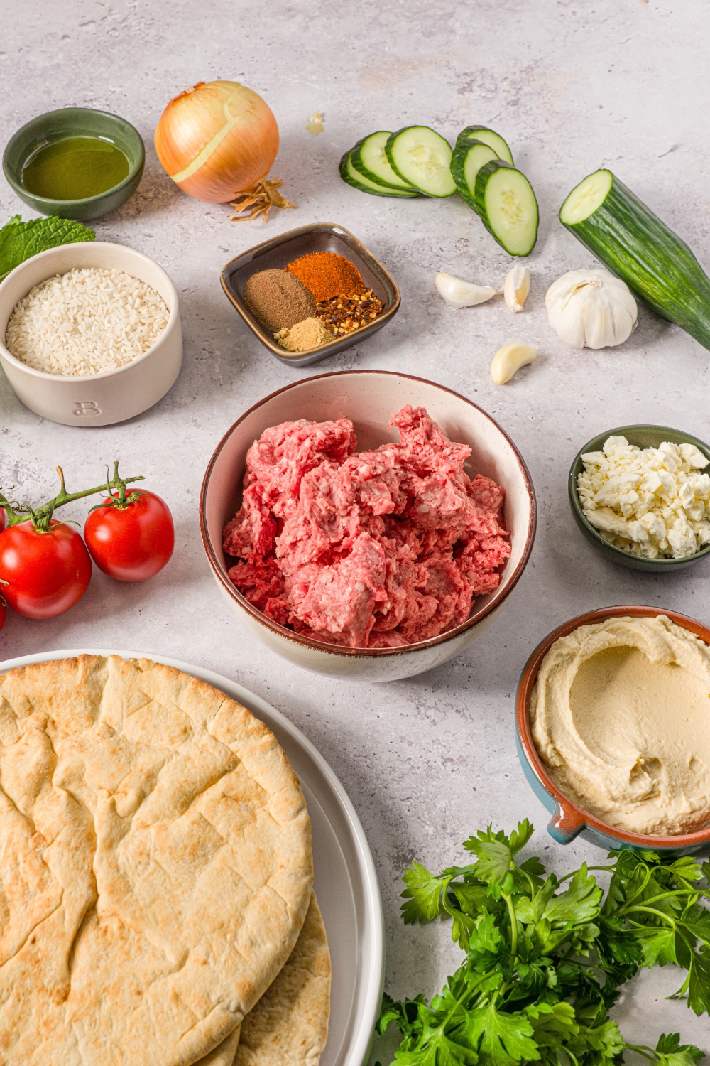 Bowls in a variety of sizes containing ingredients to make Mediterranean lamb bowls including ground lamb, garlic, cucumber, onion, feta cheese, hummus, and pita.