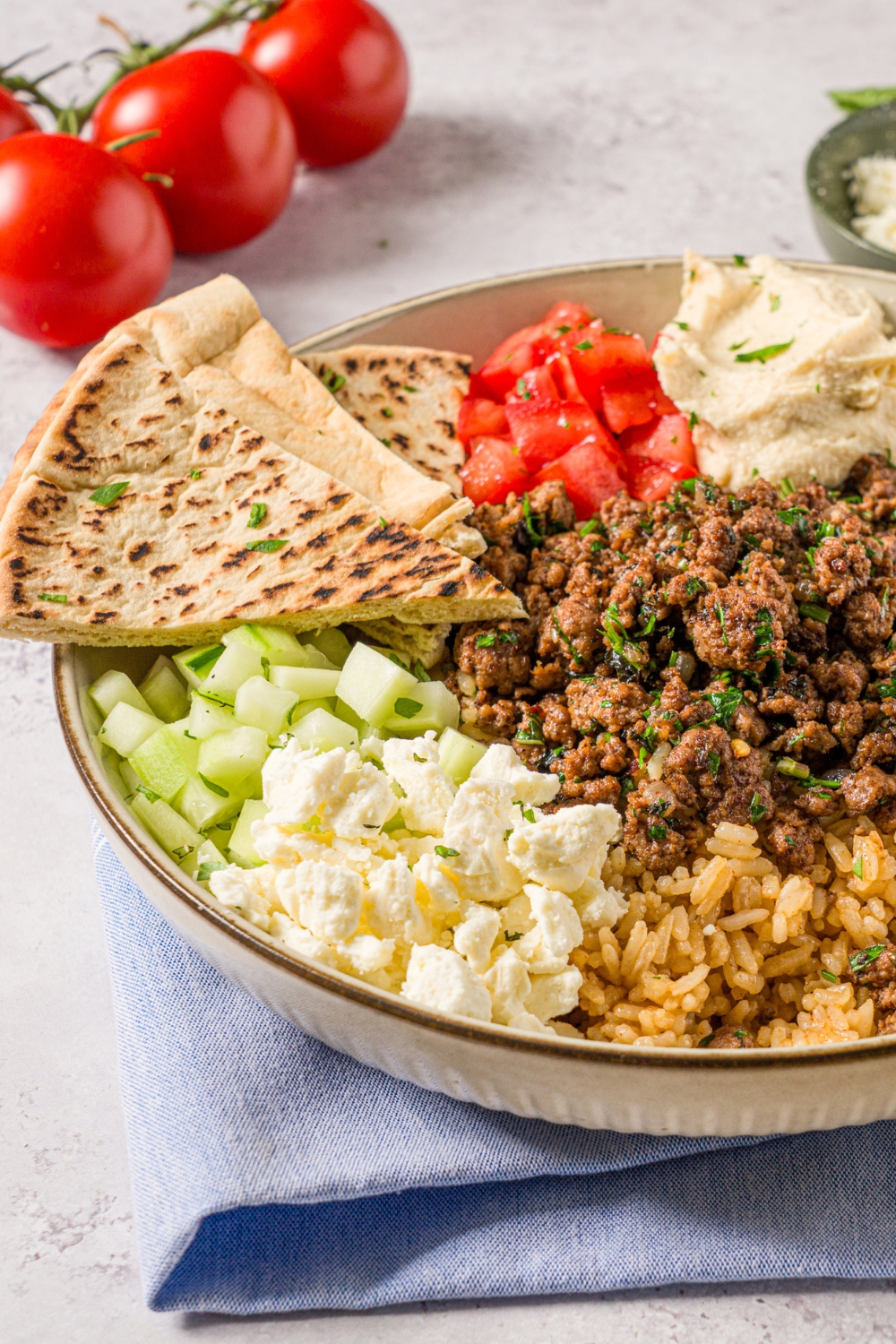 A Mediterranean lamb bowl in a ceramic bowl with brown rice, seasoned ground lamb, diced cucumbers, feta cheese, diced tomatoes, hummus, and sliced pita garnished with fresh parsley. The bowl is on a white counter with a blue cloth napkin.