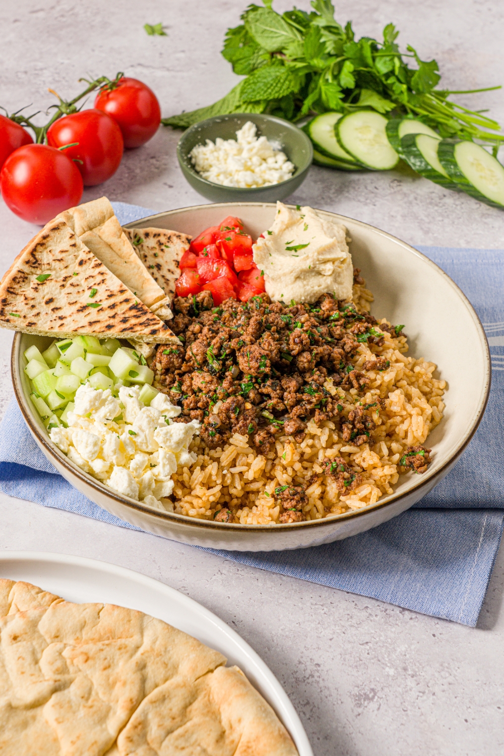 A Mediterranean lamb bowl in a ceramic bowl with brown rice, seasoned ground lamb, diced cucumbers, feta cheese, diced tomatoes, hummus, and sliced pita garnished with fresh parsley. The bowl is on a white counter with a blue cloth napkin.