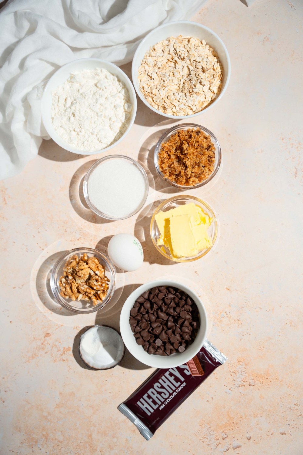 An overhead shot of several bowls in various sizes containing ingredients to make Neiman Marcus chocolate chip cookies including flour, oats, brown sugar, butter, sugar, eggs, walnuts, chocolate chips, and a Hershey's bar.
