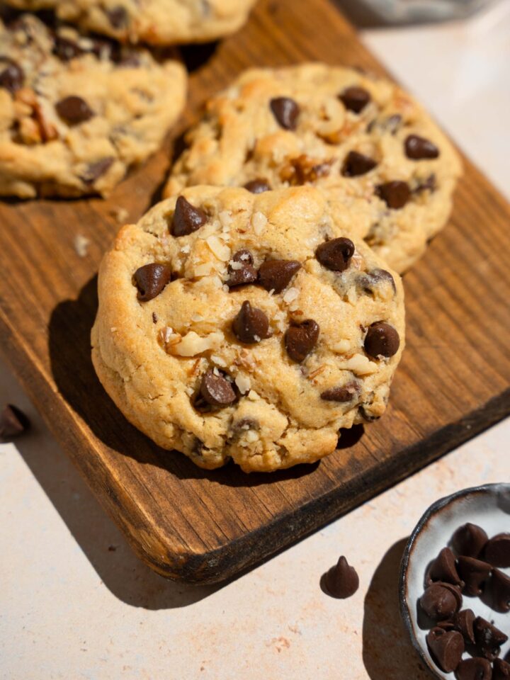 A Neiman Marcus chocolate chip cookie on a wooden board with additional cookies. The board is on a tan counter with a small plate of chocolate chips.