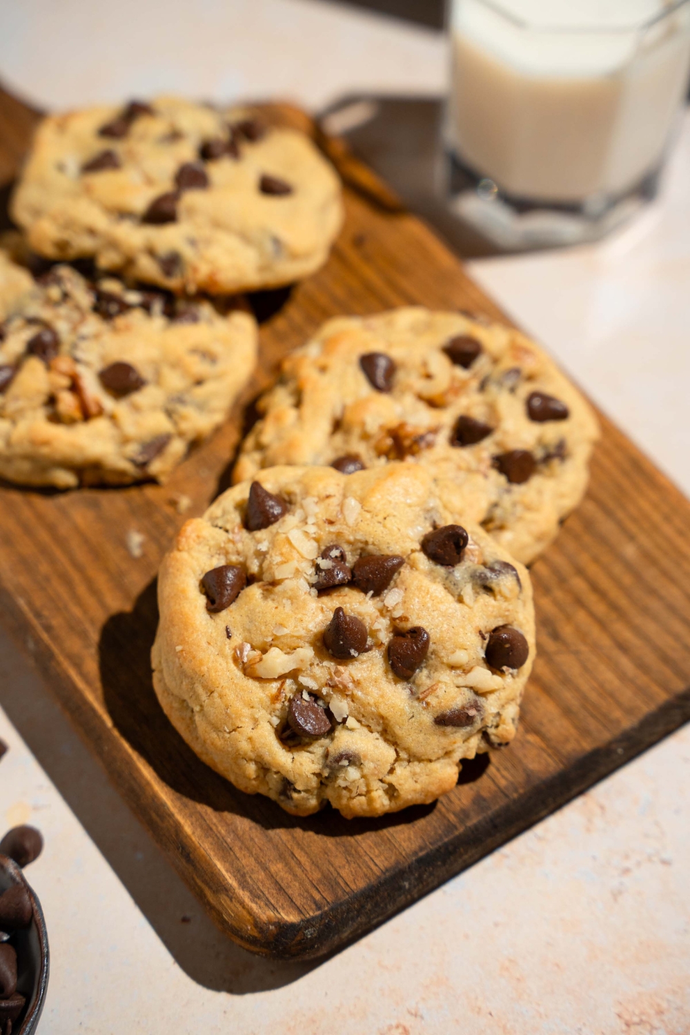 A Neiman Marcus chocolate chip cookie on a wooden board with additional cookies. The board is on a tan counter with a glass of milk.