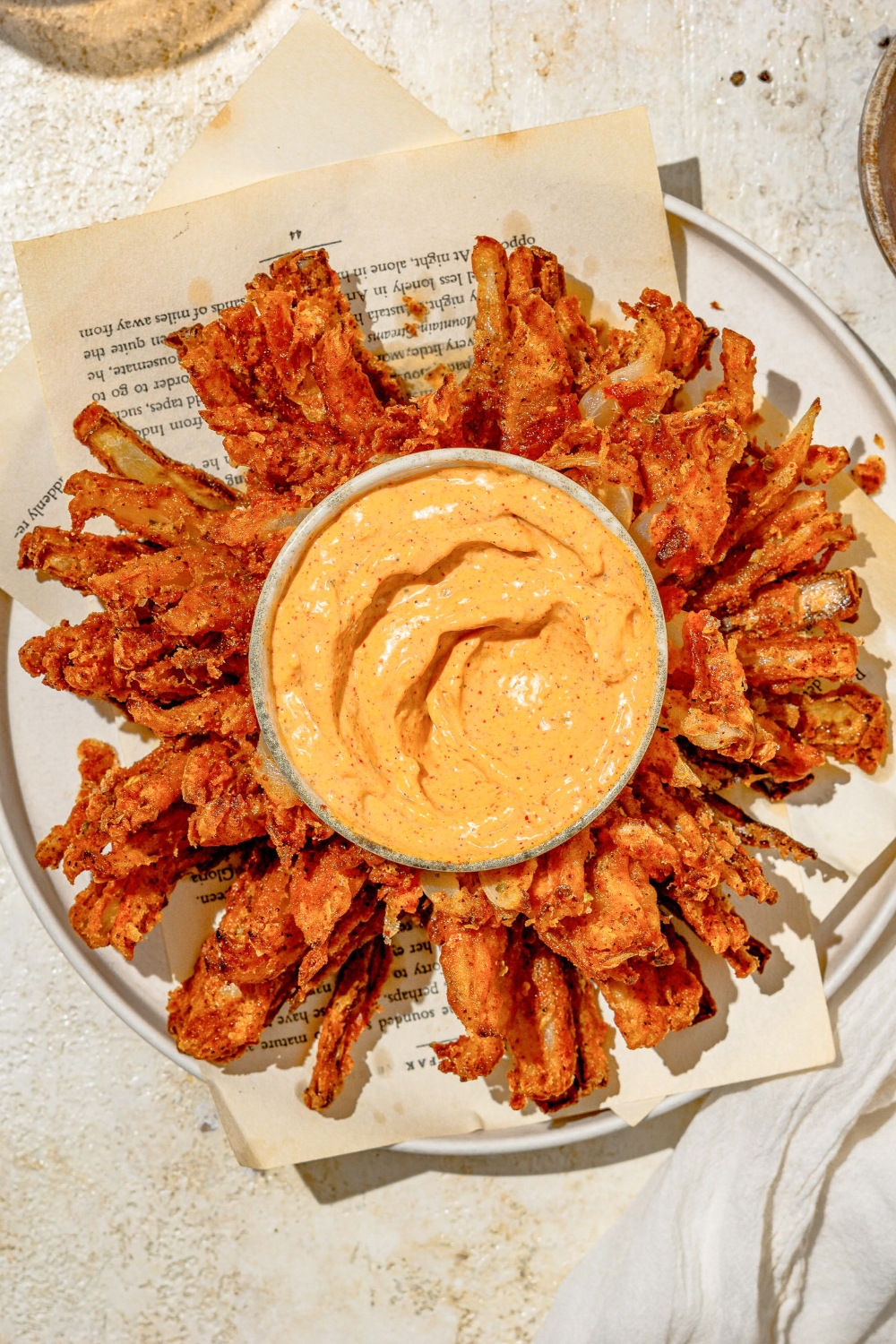 A white plate lined with paper with copycat Outback bloomin onion and a bowl of onion sauce. The plate is on a tan counter.