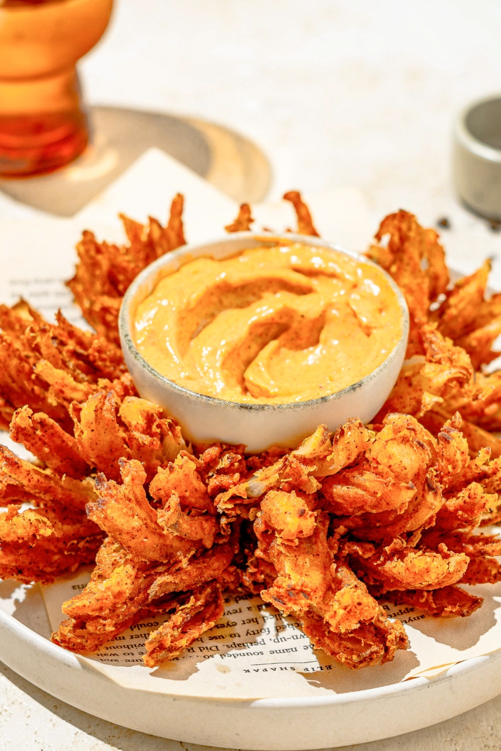 A white plate lined with paper with copycat Outback bloomin onion and a bowl of onion sauce. The plate is on a tan counter.