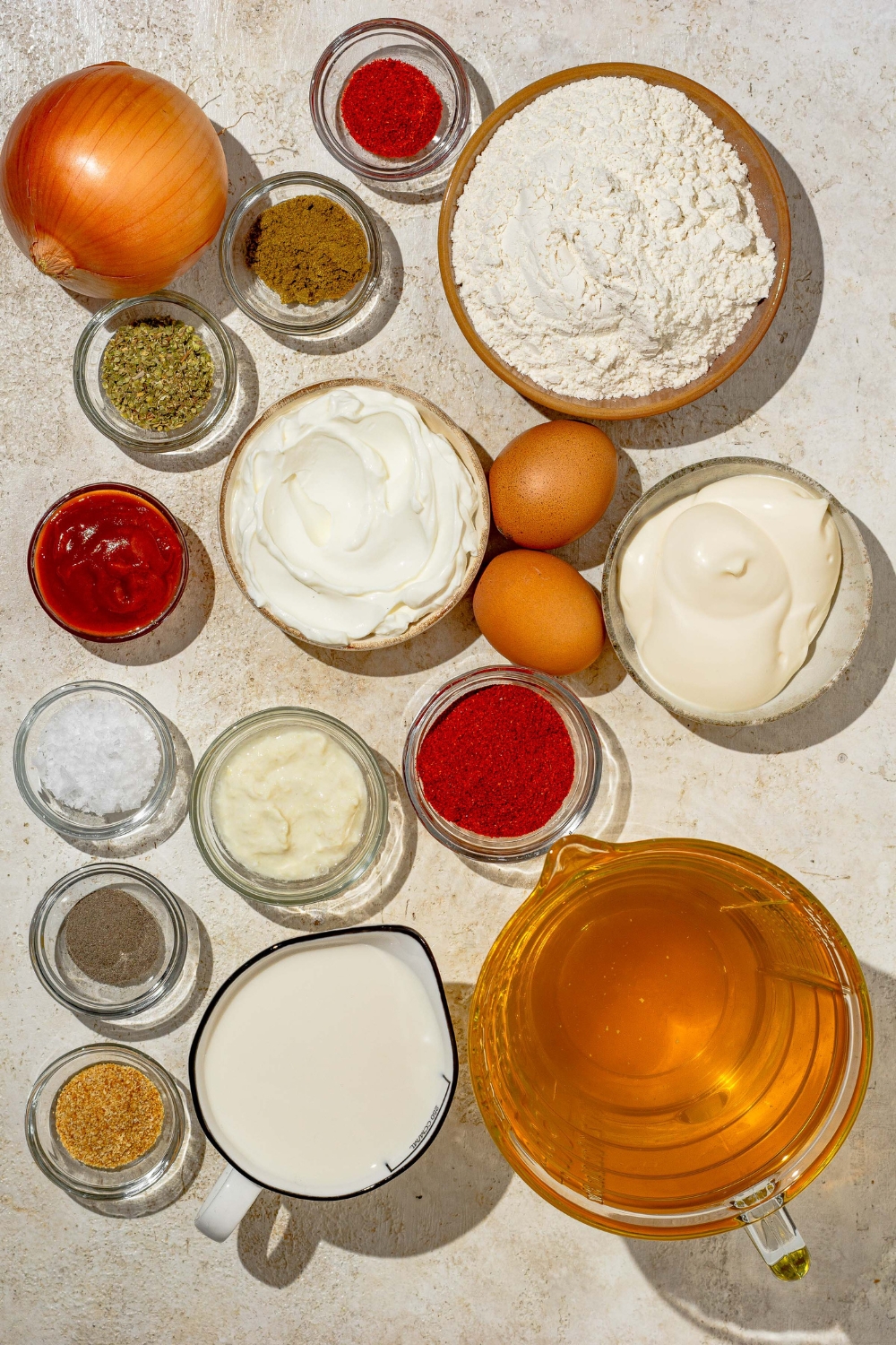 An overhead shot of several bowls of ingredients to make copycat Outback bloomin onion and bloomin onion sauce including flour, onion, eggs, mayo, sour cream, ketchup, milk, oil, and seasonings.