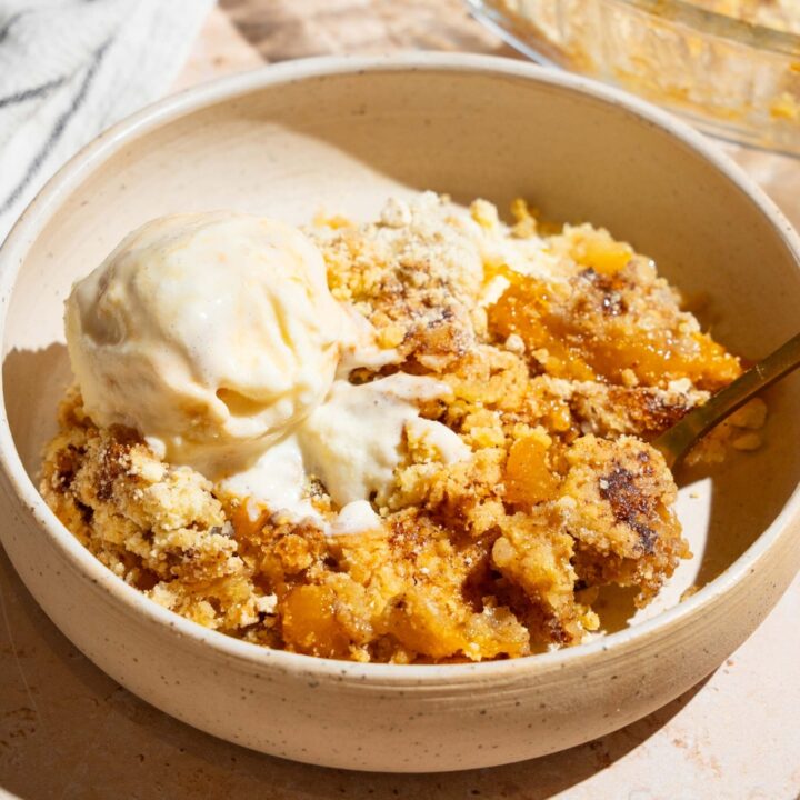 A white plate with peach cobbler dump cake topped with vanilla ice cream. There is a spoon in the plate The plate is on a tan counter with a baking dish of cake.