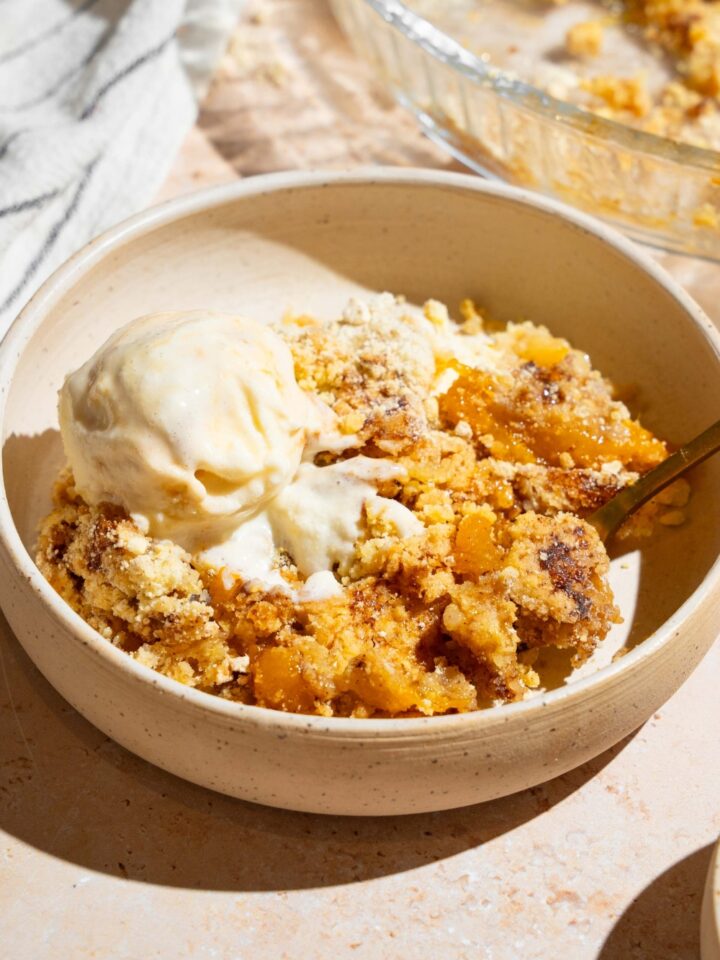A white plate with peach cobbler dump cake topped with vanilla ice cream. There is a spoon in the plate The plate is on a tan counter with a baking dish of cake.