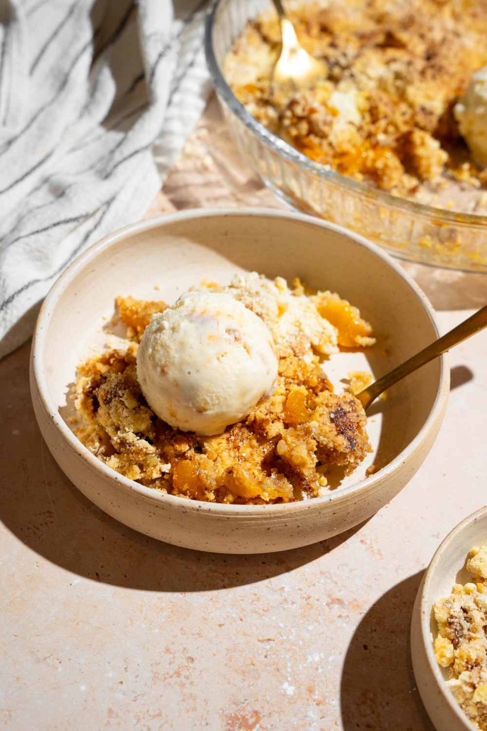 A white plate with peach cobbler dump cake topped with vanilla ice cream with a spoon. The plate is on a tan counter with a baking dish of cake.