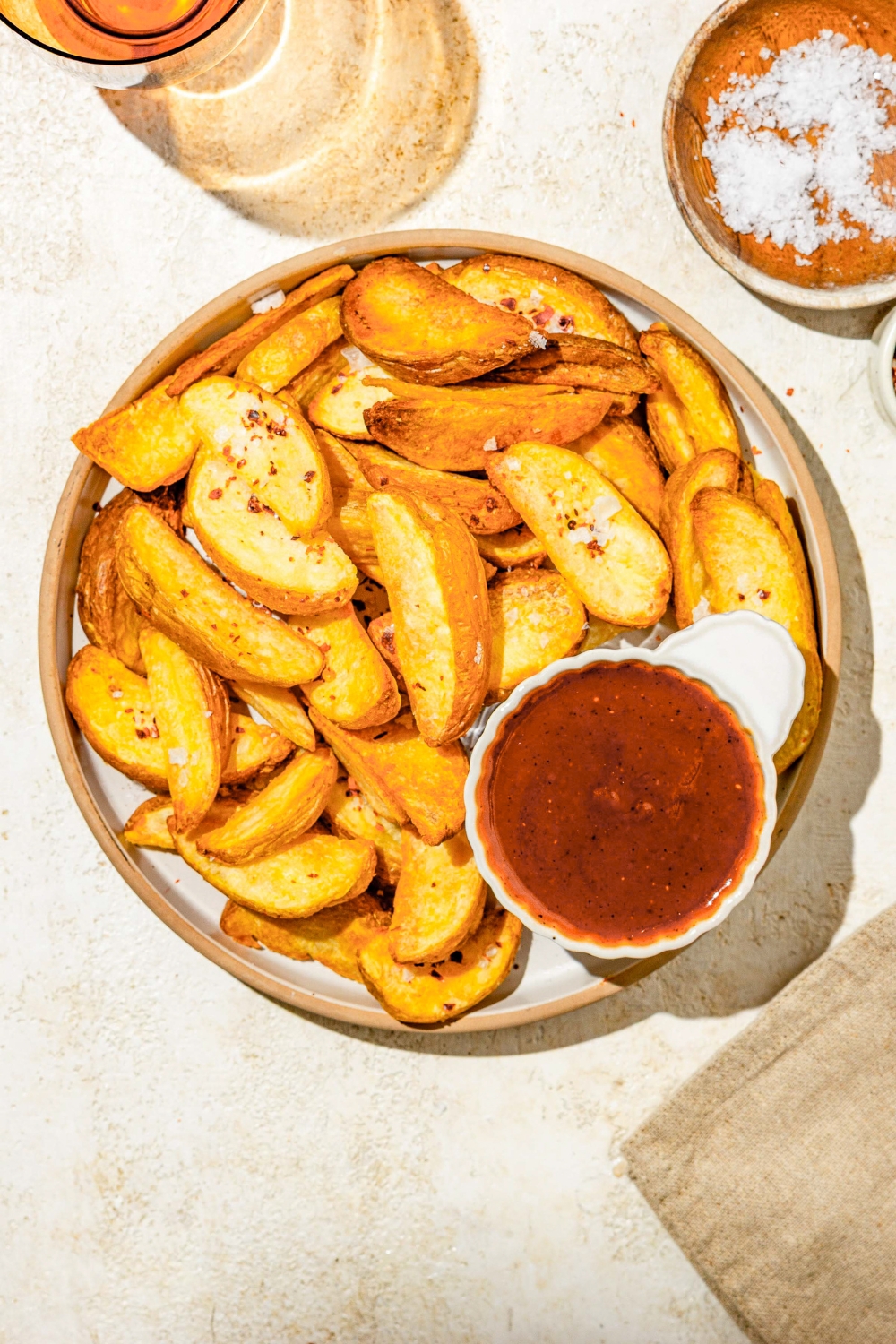 A small ramekin of Carolina BBQ sauce on plate with wedge fries. The plate is on a tan counter with a small plate of salt.