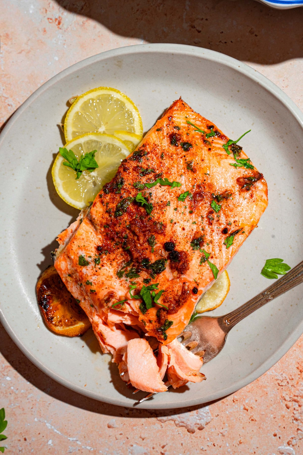 A white plate with a piece of baked Sockeye salmon garnished with fresh parsley and served with sliced lemon. There is a fork on the plate.