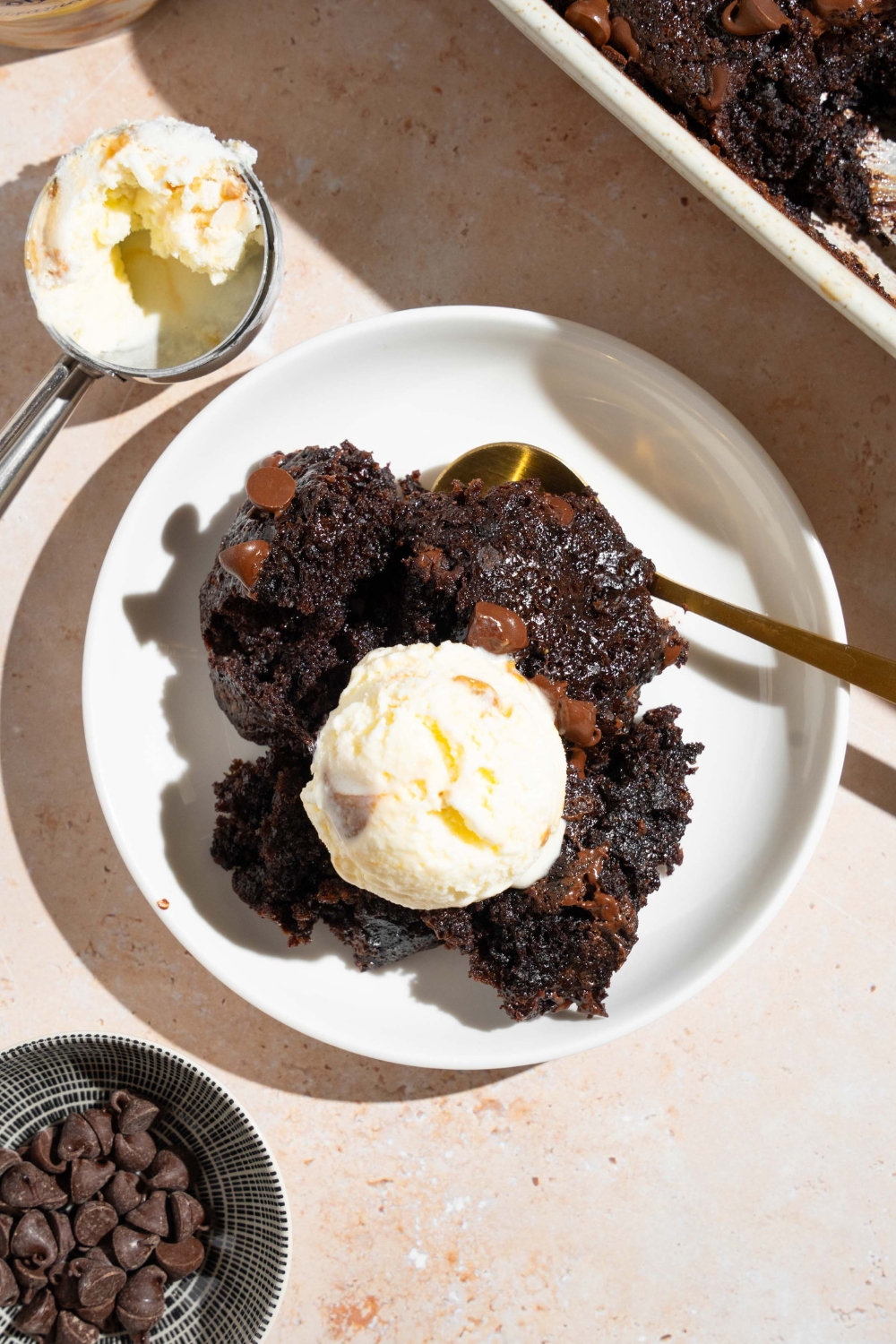A white plate with a piece of chocolate dump cake topped with vanilla ice cream. The plate is served with a spoon. The plate is on a tan counter with a baking dish of cake.