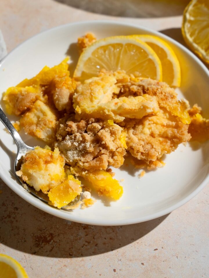 A white plate with a slice of lemon cream cheese dump cake served with sliced lemon and a fork. The plate is on a tan counter.