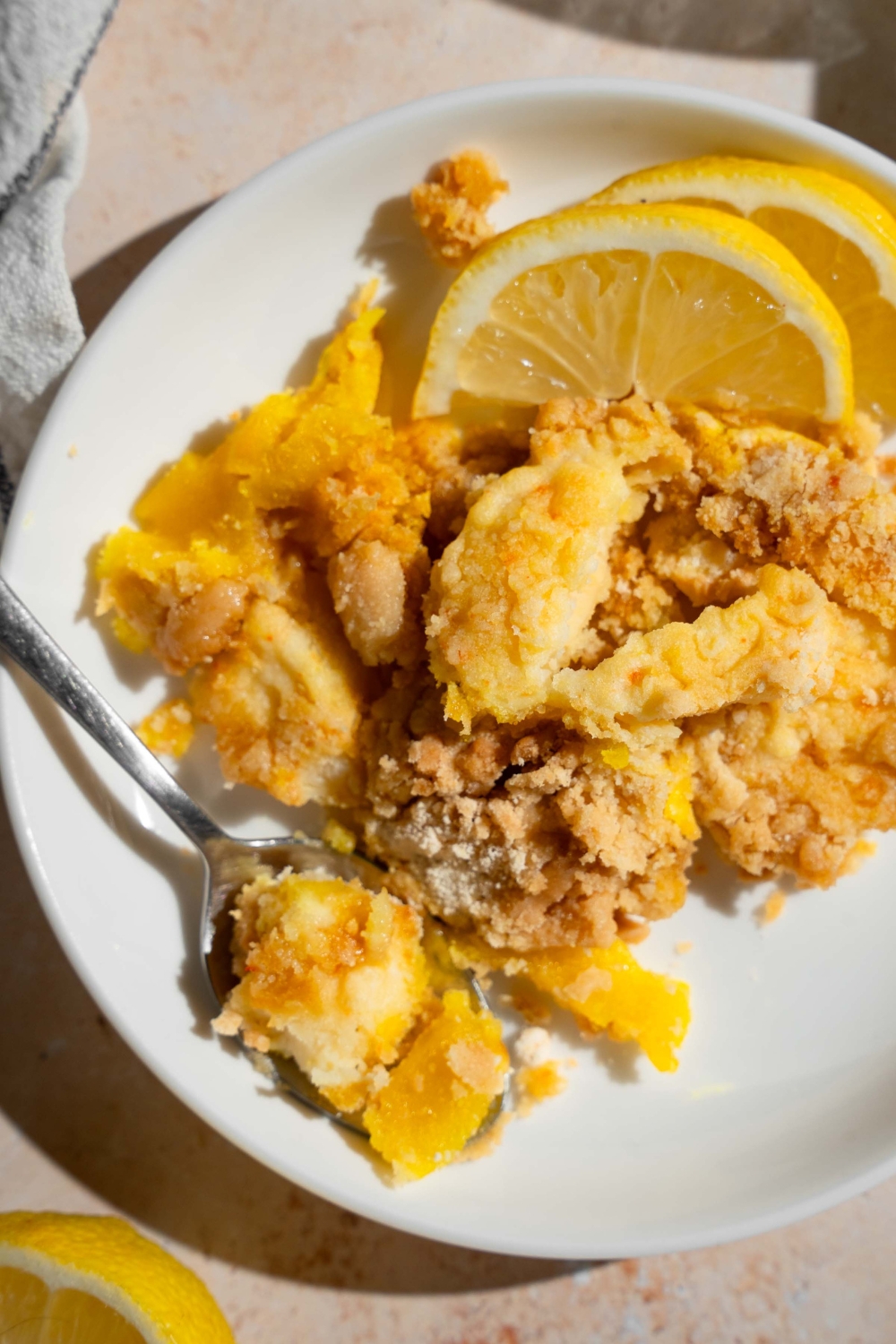 A white plate with a slice of lemon cream cheese dump cake served with sliced lemon and a fork.