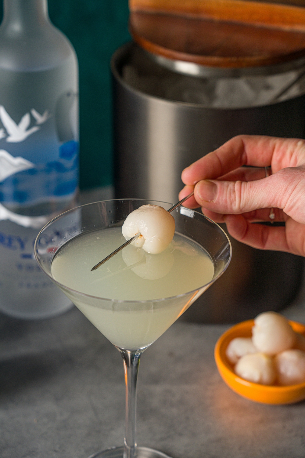 A hand placing a lychee stabbed in a toothpick into a lychee martini. The martini is on a stone counter with a small bowl of lychees, bottle of Grey Goose, and an ice bucket.