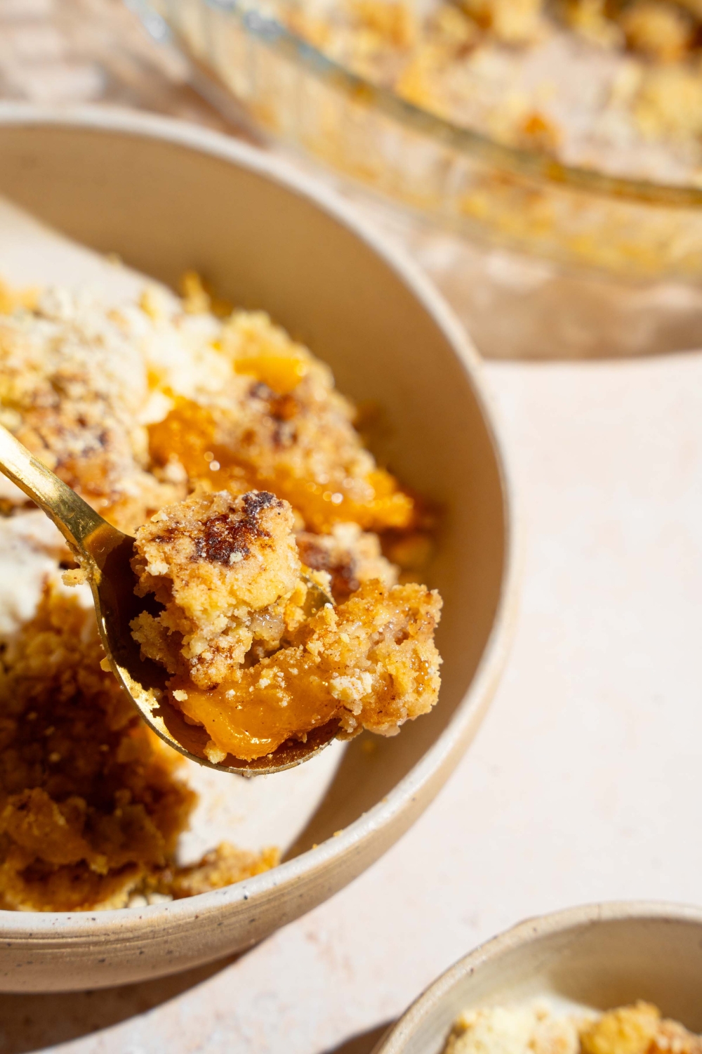 A close up of a spoon with a bite of peach cobbler dump cake. There is a bowl of cake on a tan counter blurred in the background.