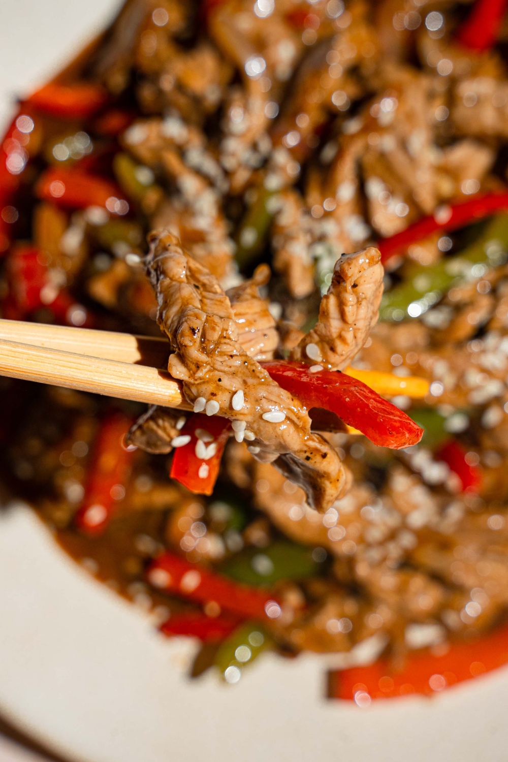 A close up of a pair of chopsticks holding a bite of pepper steak garnished with sesame seeds. There is a plate of pepper steak blurred in the background.