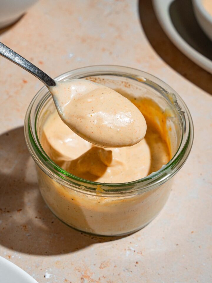A spoon dipping into a glass jar with smash burger sauce. The jar is on a tan counter.