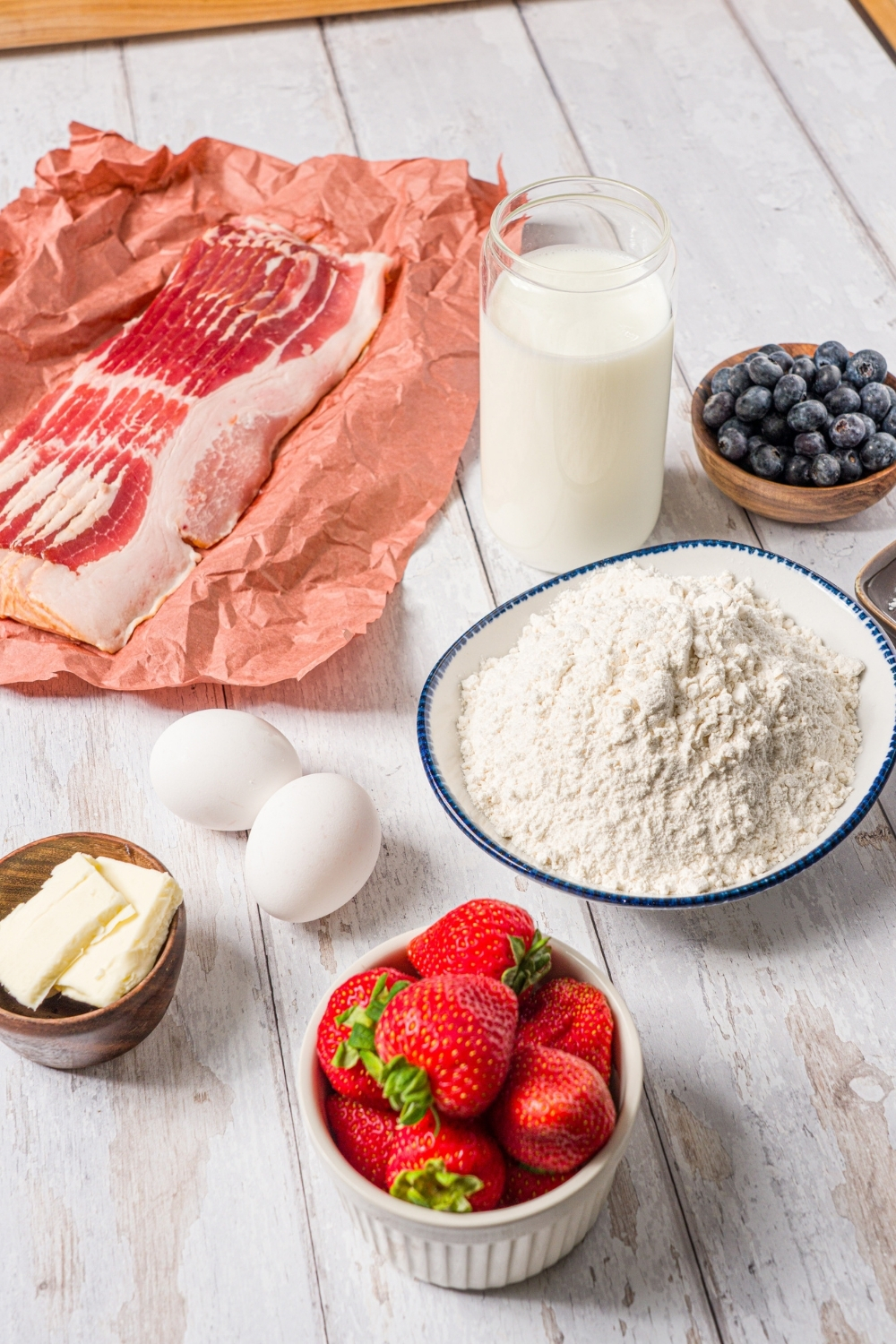 Ingredients to make reindeer pancakes on a wood counter including a uncooked bacon, flour, eggs, butter, buttermilk, blueberries, and strawberries.