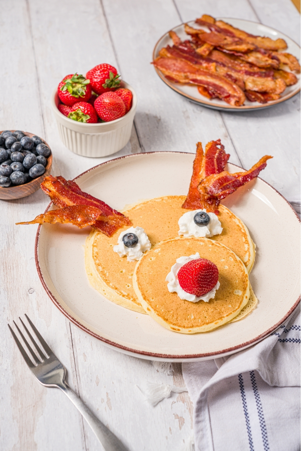 A white plate with reindeer pancakes topped with whipped cream, blueberry eyes, and a strawberry nose with bacon strips as antlers. The plate is on a wood counter with a plate of bacon and bowls of berries.