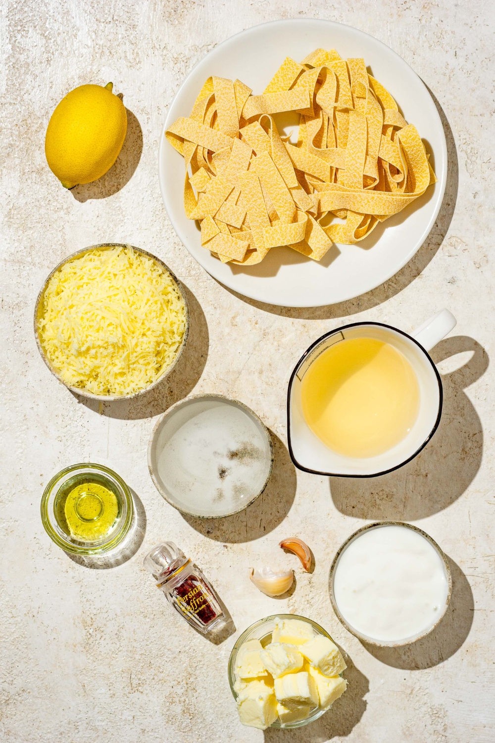An overhead shot of several bowls in various sizes containing ingredients to make saffron cream sauce including fettuccine pasta, saffron, white wine, oil, butter, garlic, parmesan cheese, and lemon.