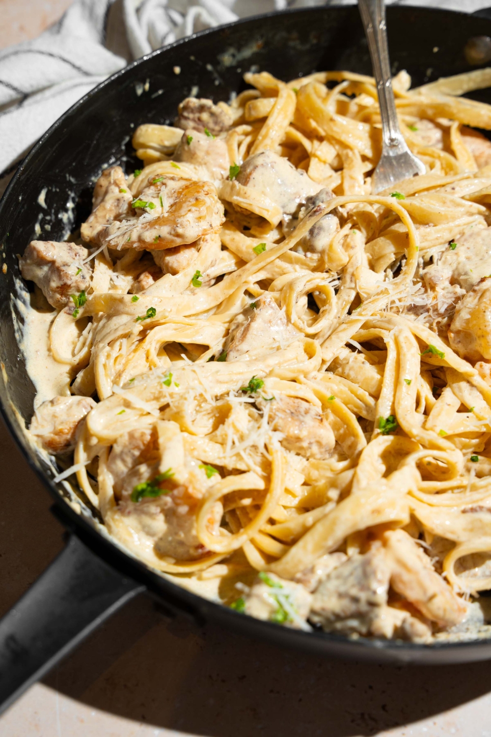 A skillet with a fork tossing chicken and shrimp alfredo garnished with fresh parsley. The skillet is on a tan counter with a white striped napkin.