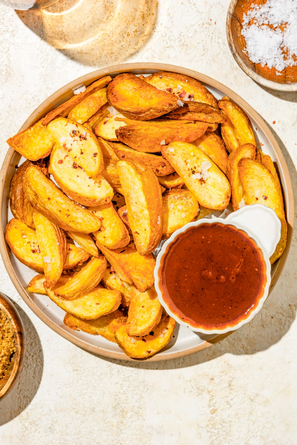A small ramekin of Carolina BBQ sauce on plate with wedge fries. The plate is on a tan counter with a small plate of salt.