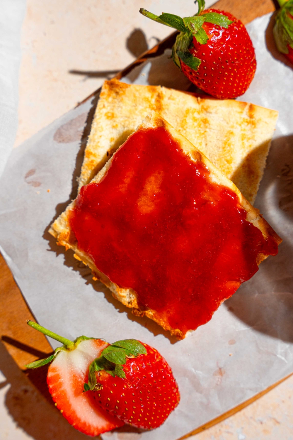 Two slices of toast with the top slice covered in Christmas jam. The toast is on a wooden board lined with parchment paper with a knife with jam and sliced strawberries. The board is on a tan counter with a jar of jam and bowl of strawberries.